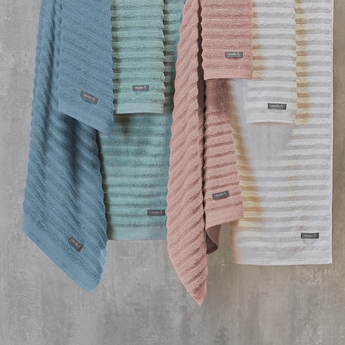 Quatro toalhas de banho com trama canelada penduradas lado a lado, em azul, verde-água, rosa e branco com listras bege