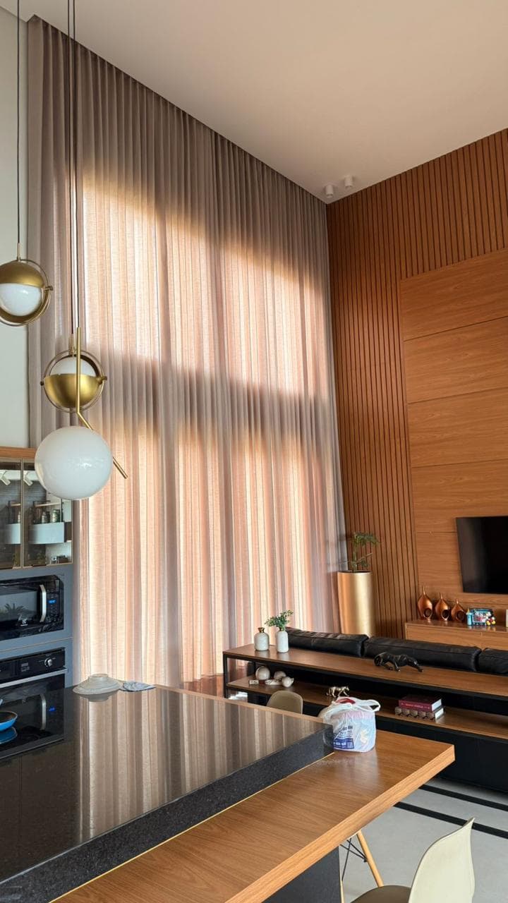 Sala integrada à cozinha, com pé-direito duplo e cortina de voil claro do teto ao chão, painel ripado de madeira e luminárias de esferas douradas