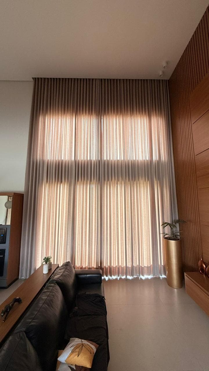 Sala de estar com pé-direito duplo, cortina de voil claro cobrindo a parede de janela inteira, sofá de couro preto e painel ripado de madeira