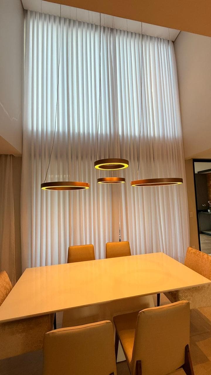 Sala de jantar com pé-direito duplo, cortina branca cobrindo a parede inteira, mesa de madeira clara, cadeiras amarelas e três luminárias circulares