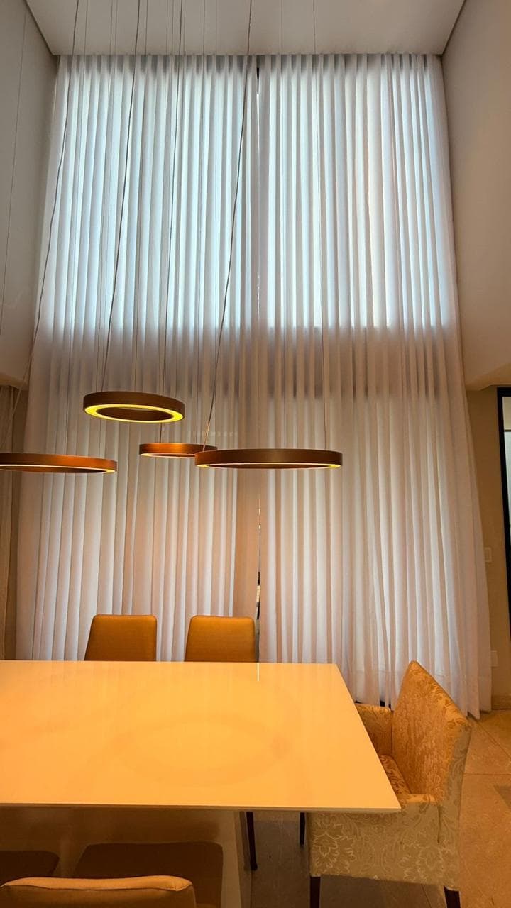 Mesa de jantar vista de perto, com a cortina branca de pé-direito duplo ao fundo filtrando a luz e luminárias circulares douradas acima