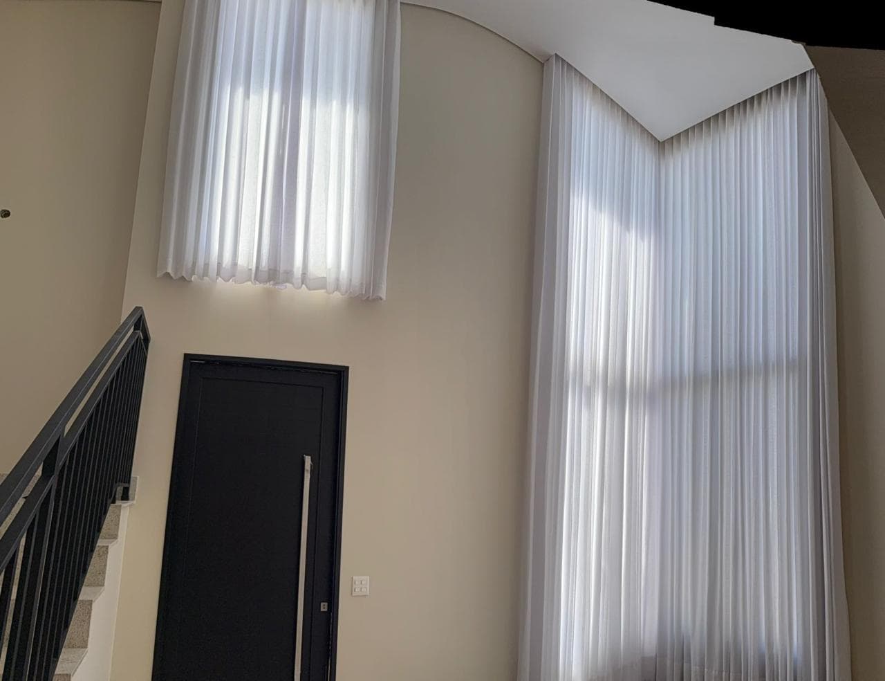 Vão de escada com pé-direito duplo, cortina branca em duas alturas acompanhando a janela, porta preta e guarda-corpo metálico