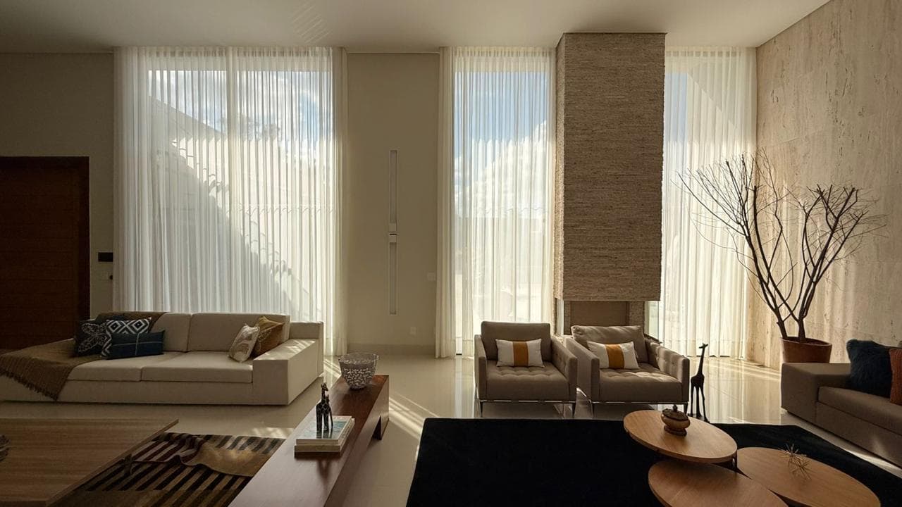 Sala ampla vista do pavimento de cima, com cortinas claras nas janelas altas, sofás bege e coluna revestida de pedra