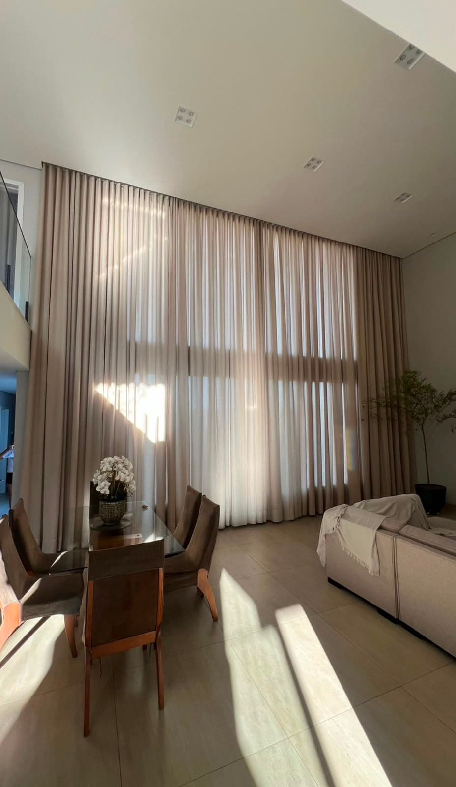 Sala de jantar integrada com pé-direito duplo, voil branco e sobrecortina bege do teto ao chão