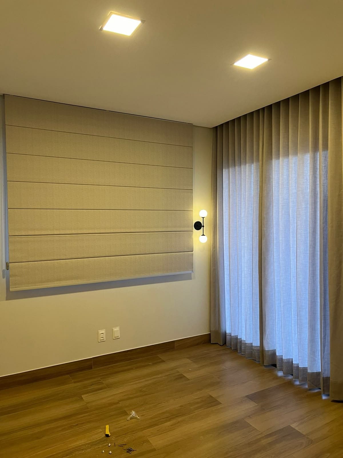 Quarto com persiana romana bege na janela e cortina de linho ao lado, piso de madeira