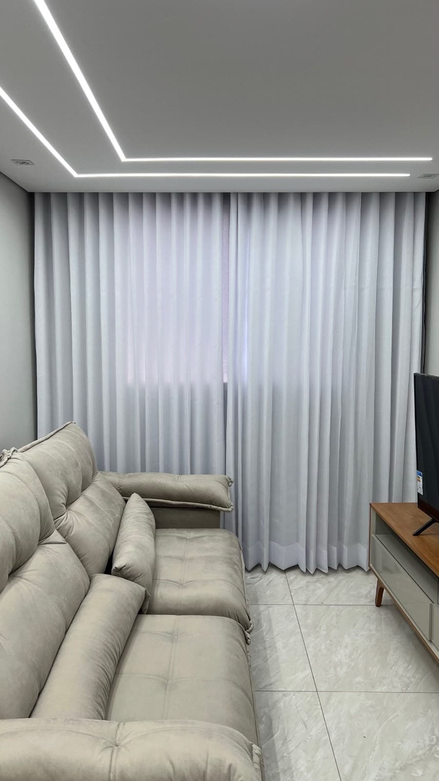 Sala de estar com cortina de voil branco e sobrecortina cinza, sofá bege e sanca com iluminação linear